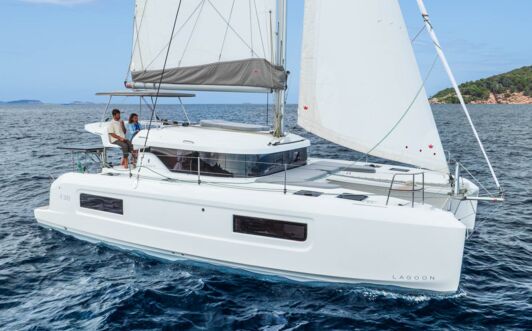Steuerbordansicht einer Lagoon 38 unter Segeln vor felsiger Küste mit Crew am Steuerstand (Werftbild) – Yachtcharter Mallorca