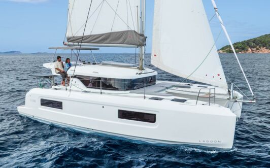 Steuerbordansicht einer Lagoon 38 unter Segeln vor felsiger Küste mit Crew am Steuerstand (Werftbild) – Yachtcharter Mallorca