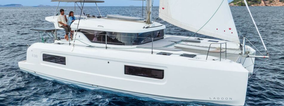Steuerbordansicht einer Lagoon 38 unter Segeln vor felsiger Küste mit Crew am Steuerstand (Werftbild) – Yachtcharter Mallorca