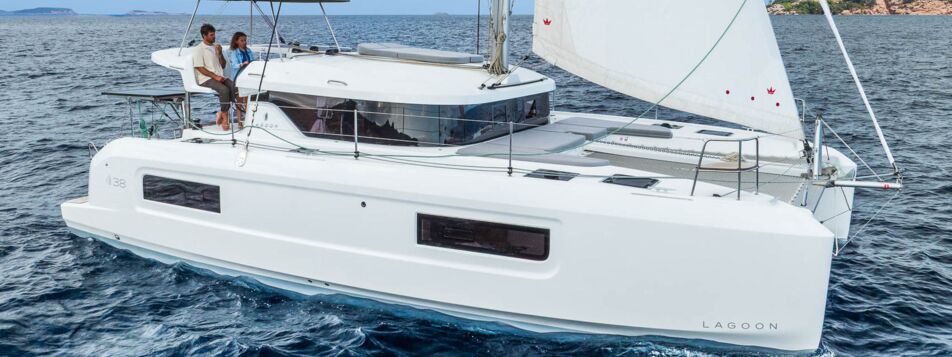 Steuerbordansicht einer Lagoon 38 unter Segeln vor felsiger Küste mit Crew am Steuerstand (Werftbild) – Yachtcharter Mallorca